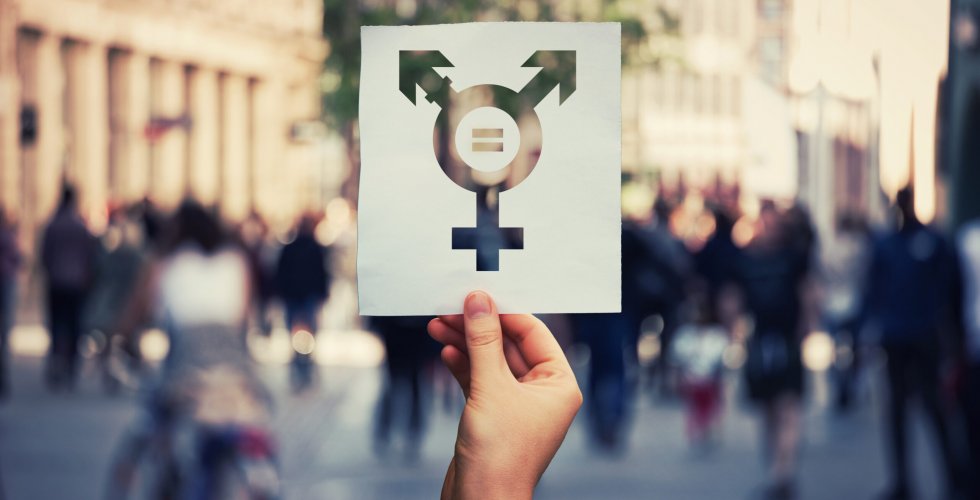 Hand holding a paper sheet with transgender symbol and equal sign inside. Equality between genders concept over a crowded city street background. Sex rights as a metaphor of social issue.